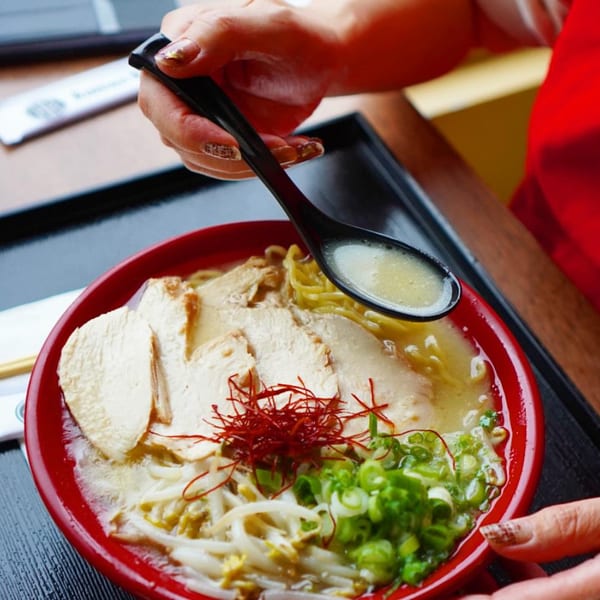 A person's hands holding a spoon over a red bowl of Ramen Ray chicken ramen.