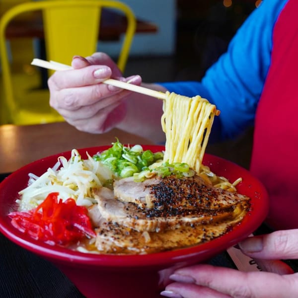 Chopsticks lifting noodles from a red bowl of Ramen Ray ramen with pork, scallions, and pickled ginger.
