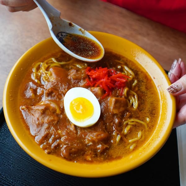 A person holding a yellow bowl of Ramen Ray curry ramen with a soft-boiled egg and red ginger.