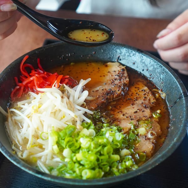 A person's hands holding a spoon with broth over a bowl of Ramen Ray ramen with toppings.
